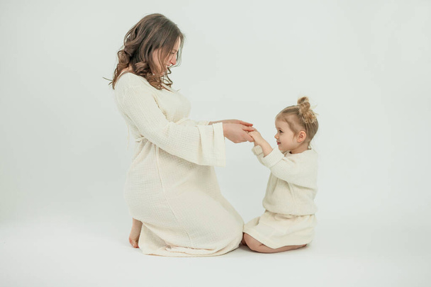 Bella giovane donna incinta con la sua piccola figlia di 3 anni su sfondo bianco. Elegante donna incinta in abito beige. Maternità. - Foto, immagini