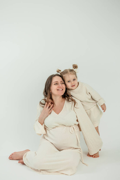 Красива молода вагітна жінка зі своєю маленькою 3-річною дочкою на білому тлі. Стильна вагітна жінка в бежевій сукні. Батьківство. - Фото, зображення