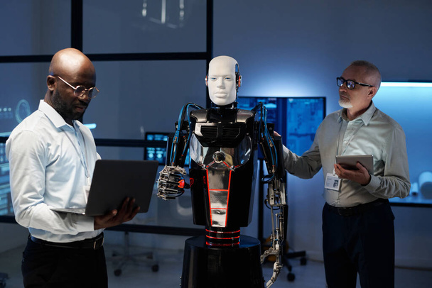 Μηχανικοί που χρησιμοποιούν gadgets για να συνδέσουν ρομπότ κατά τη διάρκεια της ομαδικής εργασίας τους στο εργαστήριο - Φωτογραφία, εικόνα