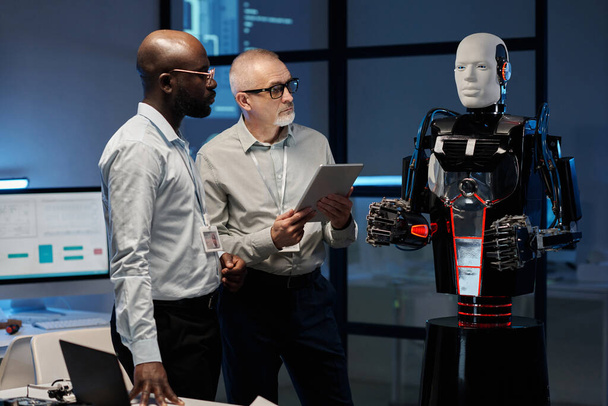 Δύο πολυεθνικοί μηχανικοί εξετάζουν το πρόγραμμα των ρομπότ χρησιμοποιώντας ψηφιακή ταμπλέτα ενώ στέκονται στο γραφείο - Φωτογραφία, εικόνα