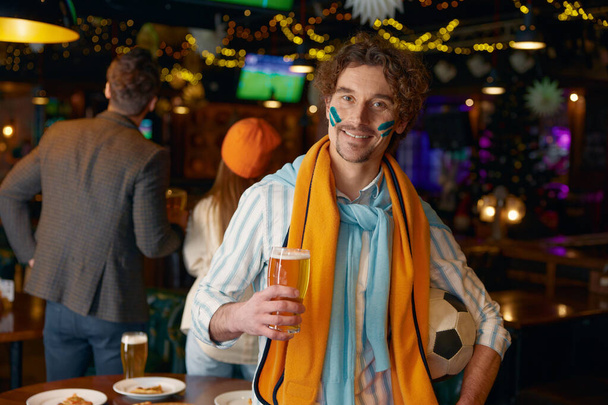 Retrato de chico guapo de fútbol sonriendo a la cámara sosteniendo la pelota de fútbol y vaso de cerveza espumosa bebida de pie sobre el fondo interior del bar deportivo - Foto, Imagen