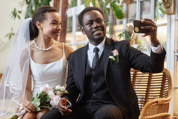 Portrait of young black couple as bride and groom taking selfie photo together in green orangerie - Photo, Image