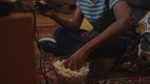 Inclinazione filmati palmari di afro-americano gen Y bambino mangiare una manciata di popcorn durante la riproduzione di videogiochi console davanti alla TV seduto sul pavimento in stile retrò soggiorno, atmosfera degli anni '90 - Filmati, video