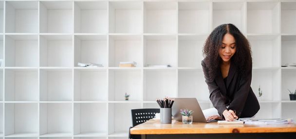 Femme professionnelle se penche sur son bureau, intensément axée sur l'écriture de notes avec un ordinateur portable ouvert devant elle dans un bureau moderne. - Photo, image