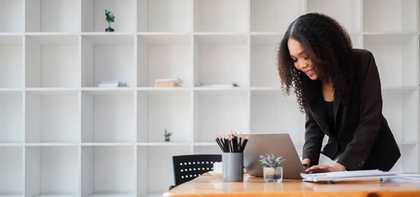 Empresária concentrada está ocupada no trabalho, em pé sobre sua mesa com documentos e um laptop em um ambiente de escritório moderno. - Foto, Imagem