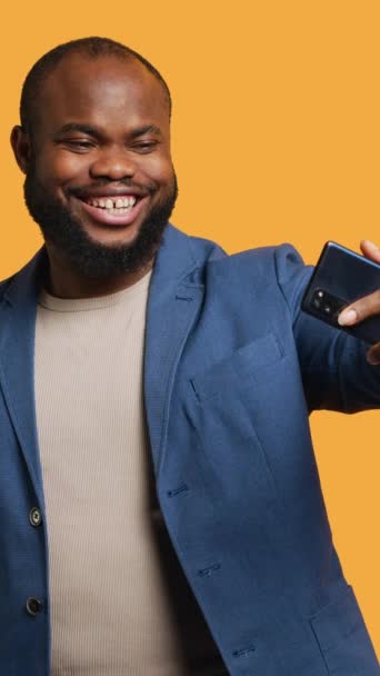 Вертикальне відео Joyous людина, яка використовує смартфон, щоб взяти селфі і розмістити їх у соціальних мережах. Щаслива людина BIPOC робить фотографії за допомогою камери селфі телефону, ізольовані на жовтому студійному фоні, камера А - Кадри, відео
