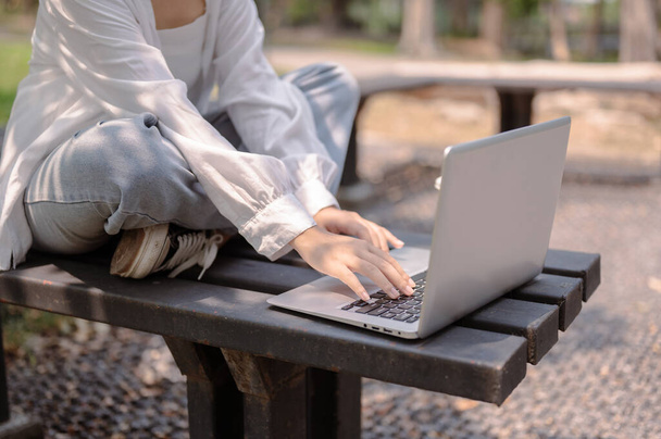 Μια κουρεμένη εικόνα Ασιάτισσας με καθημερινά ρούχα κάθεται σε ένα παγκάκι σε δημόσιο πάρκο και χρησιμοποιεί τον φορητό υπολογιστή της, δουλεύοντας εξ αποστάσεως σε εξωτερικούς χώρους μια ηλιόλουστη μέρα. ιδέες τρόπου ζωής και ασύρματης τεχνολογίας - Φωτογραφία, εικόνα