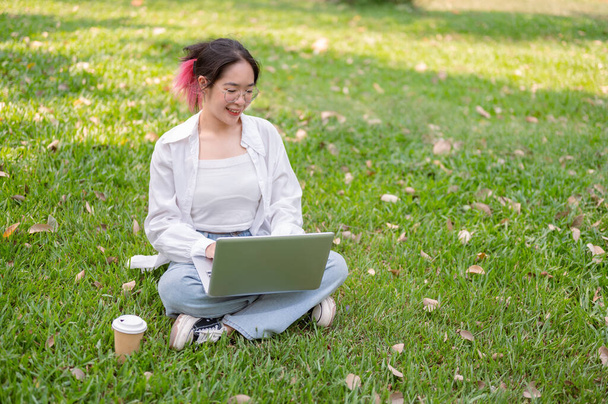 Молодая счастливая азиатка в очках и повседневной одежде сидит на траве в парке, работая над своим ноутбуком. удаленная работа, беспроводные технологии, современный городской образ жизни - Фото, изображение