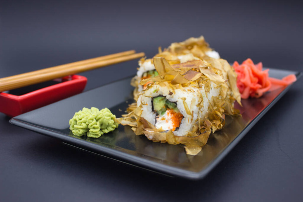 Fotografia przedstawia wykwintne ustawienie sushi, wraz z rolką zwieńczoną delikatnymi płatkami bonito, spoczywającymi na czarnym prostokątnym talerzu. Towarzyszą sushi są żywe zielone wasabi i kopiec marynowanego imbiru, z parą bambusa cho - Zdjęcie, obraz