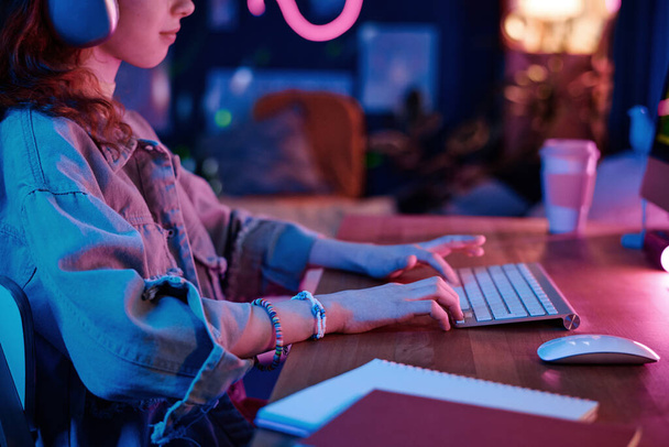 Widok z boku ujęcie nowoczesnej nastolatki w słuchawki siedzi przy biurku gry wideo na komputerze w jej pokoju wieczorem - Zdjęcie, obraz