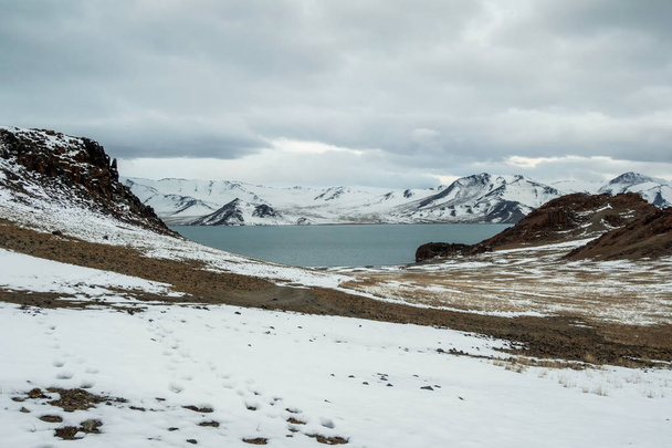 Грязная дорога к озеру покрыта снегом. Панорамный вид на большое озеро в облачное утро. Монгольские природные ландшафты вблизи озера Толбо-Нуур, окруженные горами и скалами на севере Монголии. - Фото, изображение