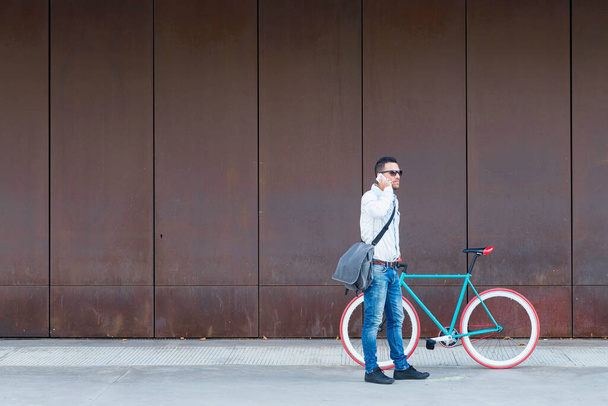 Stylish man with sunglasses and a messenger bag standing by a red-wheeled bike, making a phone call against a rusted metal wall - Photo, Image