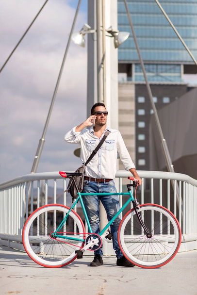 Modieuze man in wit hemd en zonnebril staat met een kleurrijke fiets op een stadsbrug, hoogbouw op de achtergrond - Foto, afbeelding