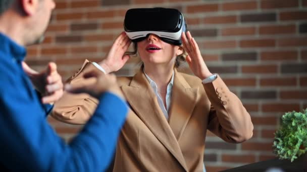 Женщина впечатлена использованием гарнитуры Virtual Reality в офисе, в то время как мужчина объясняет и жестикулирует - Кадры, видео