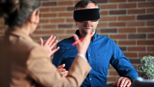 Homme utilisant un casque de réalité virtuelle dans un bureau pendant que la femme explique et gesticule - Séquence, vidéo