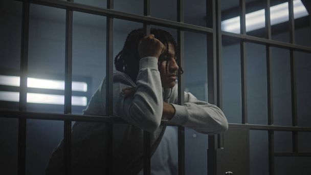 Upset African American teenager with face tattoos stands in prison cell in jail or youth detention center leaning on metal bars. Prison officer passes by young criminal or prisoner in the foreground. - Photo, Image