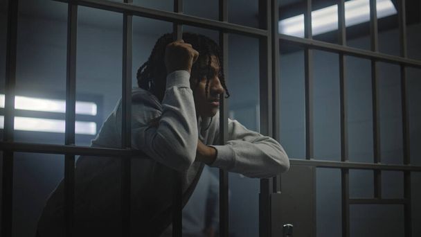 Zdenerwowany afro-amerykański nastolatek z tatuażami na twarzy stoi w więziennej celi lub w ośrodku dla młodzieży opierającym się o metalowe kraty. Oficer więzienny przechodzi przez młodego przestępcę lub więźnia na pierwszym planie. - Zdjęcie, obraz