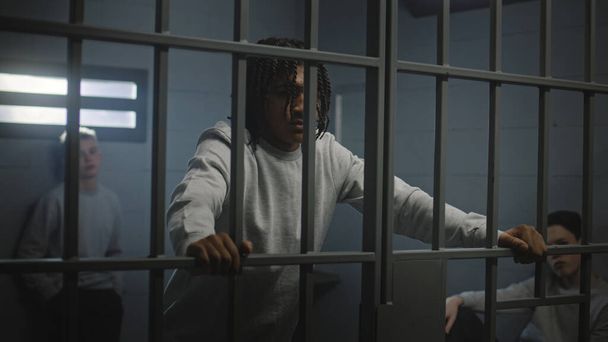 Adolescentes multiétnicos tras las rejas de metal en la celda de la prisión miran a la cámara. Jóvenes prisioneros, criminales cumplen penas de prisión por crímenes en la cárcel. Centro de detención juvenil o correccional. Retrato - Foto, imagen