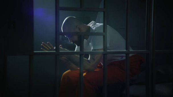 Criminal in orange uniform sits on bed in prison cell, stands up and looks at barred window. Prisoner serves imprisonment term for crime in jail. Gangster in detention center. View through metal bars. - Photo, Image