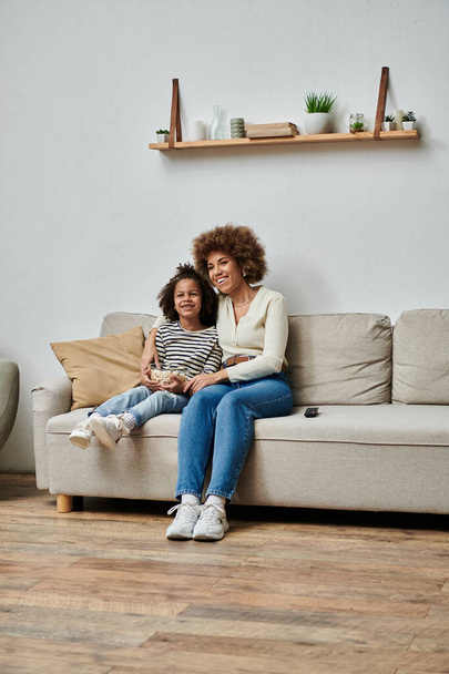 Μια ευτυχισμένη Αφρο-Αμερικανίδα μητέρα και κόρη κάθονται σε έναν καναπέ σε ένα άνετο σαλόνι, χαμογελώντας και απολαμβάνοντας ο ένας την παρέα του άλλου. - Φωτογραφία, εικόνα