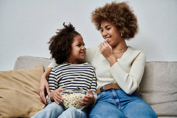 Feliz madre afroamericana y su hija se sientan en un sofá, disfrutando de un bocadillo de palomitas de maíz juntas. - Foto, imagen