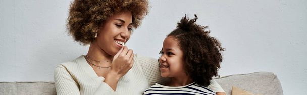 Афроамериканская мать и дочь делятся улыбками, сидя на уютном диване, наслаждаясь временем вместе дома. - Фото, изображение