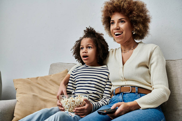 Happy African American mother and daughter sit together on a couch, watching TV with smiles on their faces. - Photo, Image