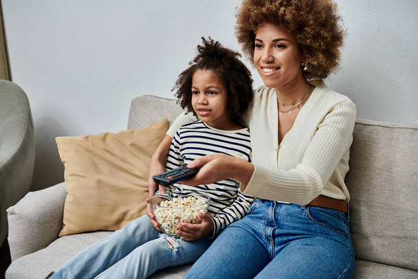 Любящие афро-американская мать и дочь счастливо сидят на диване и смотрят телевизор вместе.. - Фото, изображение