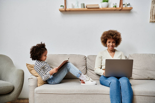 Una madre e una figlia afroamericana felicemente sedute su un divano, assorte nell'uso di un computer portatile insieme. - Foto, immagini