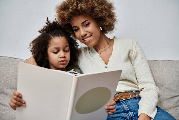Eine glückliche afroamerikanische Mutter und Tochter sitzen auf einer Couch, in ein Buch vertieft und teilen einen besonderen Moment miteinander. - Foto, Bild