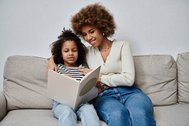 Afro-Amerikalı neşeli bir anne ve kız kanepede rahat bir şekilde otururken büyüleyici bir kitaba daldılar.. - Fotoğraf, Görsel