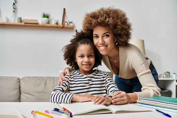 Una feliz madre afroamericana y su hija se sientan juntas en una mesa, disfrutando de un tiempo de calidad en casa. - Foto, Imagen