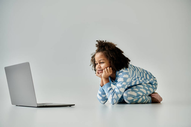A happy African American girl in pajamas sits on the floor engrossed in a laptop - Фото, зображення