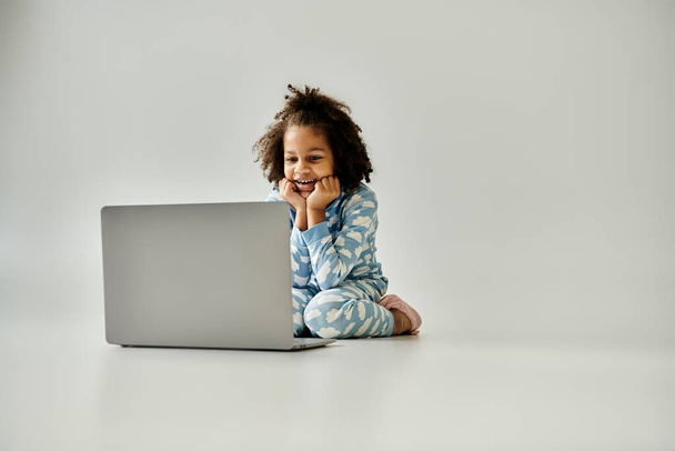 パジャマの若いアフリカ系アメリカ人の少女がノートパソコンで床に座り,デジタル世界を探検する. - 写真・画像