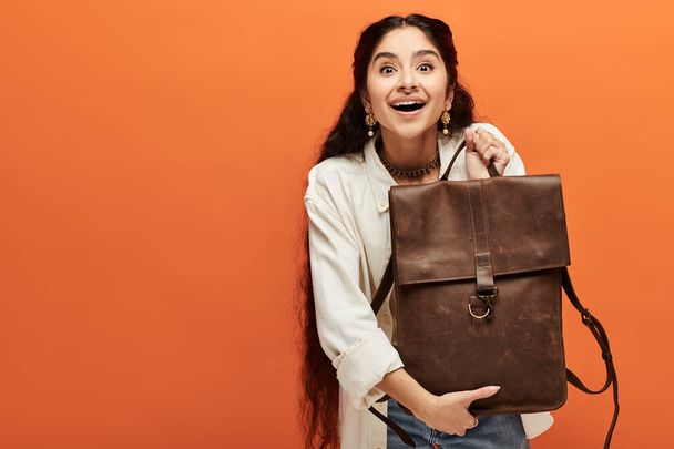 Genç Hintli kadın açık turuncu bir arka planda şık kahverengi bir sırt çantası sergiliyor.. - Fotoğraf, Görsel