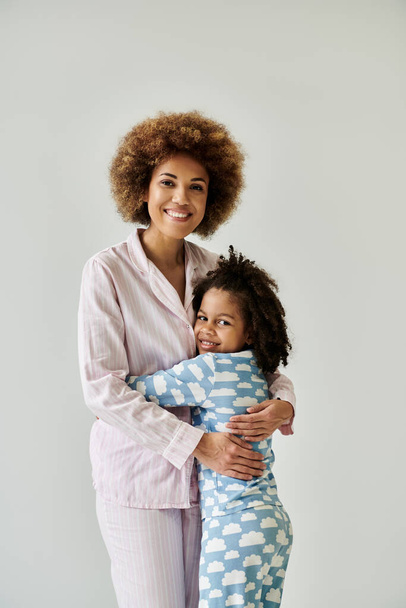 Χαρούμενη Αφρο-Αμερικανίδα μητέρα και κόρη μοιράζονται μια ζεστή αγκαλιά με ζεστές πιτζάμες σε γκρι φόντο. - Φωτογραφία, εικόνα