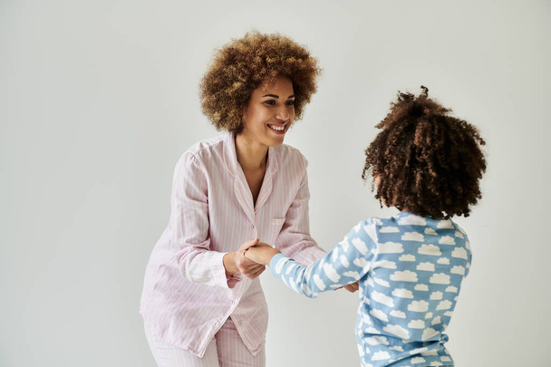 Μια Αφρο-Αμερικανίδα μητέρα και κόρη με πιτζάμες κάνουν χειραψία, ακτινοβολώντας ζεστασιά και ενότητα σε ένα γκρίζο φόντο.. - Φωτογραφία, εικόνα