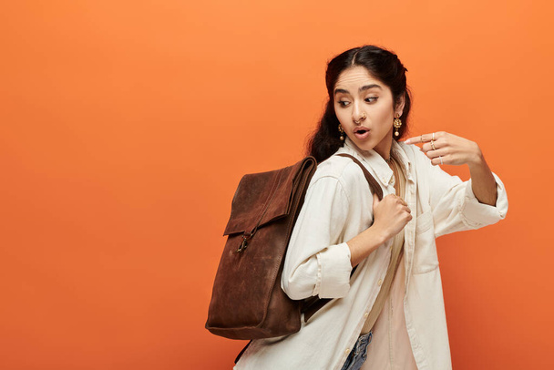 茶色のバックパックを持つ若いインドの女性は鮮やかなオレンジ色の背景にポーズします. - 写真・画像