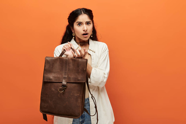 Young indian woman energetically posing with a brown backpack against vibrant orange background. - Photo, Image