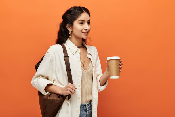 Μια νεαρή Ινδή γυναίκα κρατάει ένα φλιτζάνι καφέ πάνω σε ένα φωτεινό πορτοκαλί φόντο.. - Φωτογραφία, εικόνα