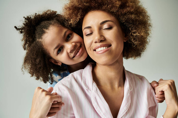 Sonriendo madre e hija afroamericana en pijama, posando felizmente sobre un fondo gris. - Foto, Imagen
