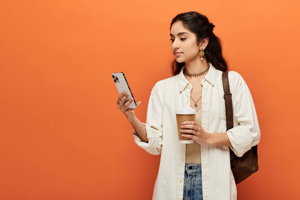 Een jonge indiaanse vrouw houdt een kopje koffie terwijl een blik op haar telefoon scherm. - Foto, afbeelding