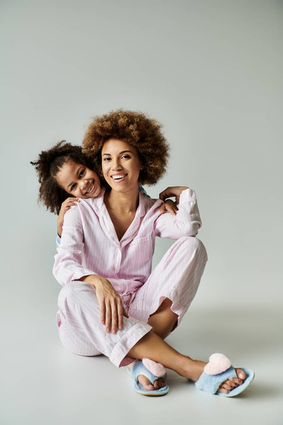 Uma mãe e filha afro-americana, vestida de pijama, posando alegremente para um retrato elegante em um fundo cinza. - Foto, Imagem
