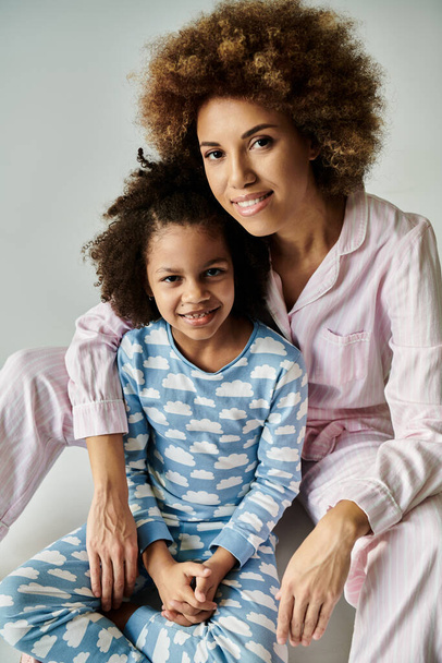 Lächelnde afroamerikanische Mutter und Tochter posieren in bunten Pyjamas vor sanftem grauen Hintergrund. - Foto, Bild