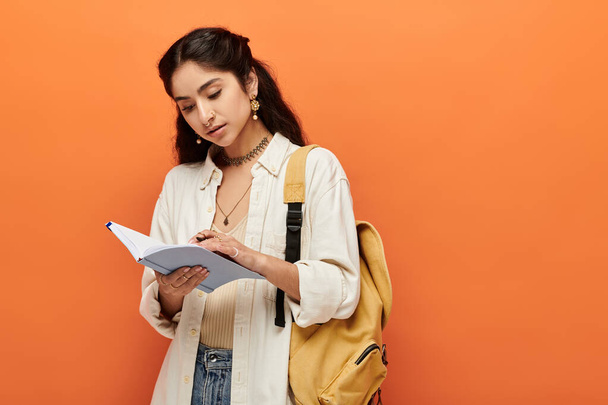 Young indian woman actively writing in notebook against vibrant orange background. - Photo, Image