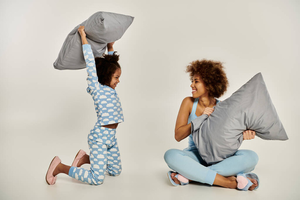 Szczęśliwa Afroamerykanka matka i córka w piżamie angażują się w zabawną walkę na poduszce na szarym tle. - Zdjęcie, obraz