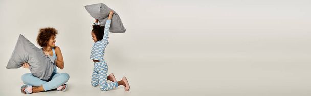 Gioioso afroamericano madre e figlia in pigiama giocosamente gettando cuscini in un momento accogliente su uno sfondo grigio. - Foto, immagini