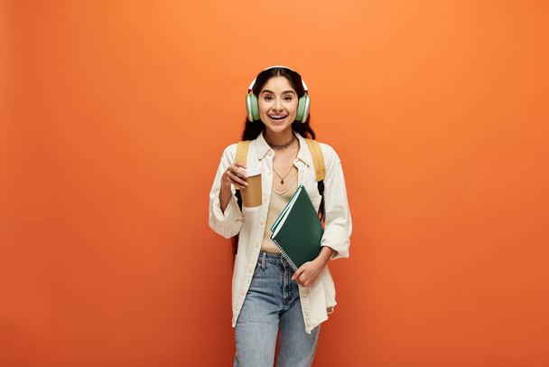 Young indian woman wearing headphones holds book against orange backdrop. - Photo, Image