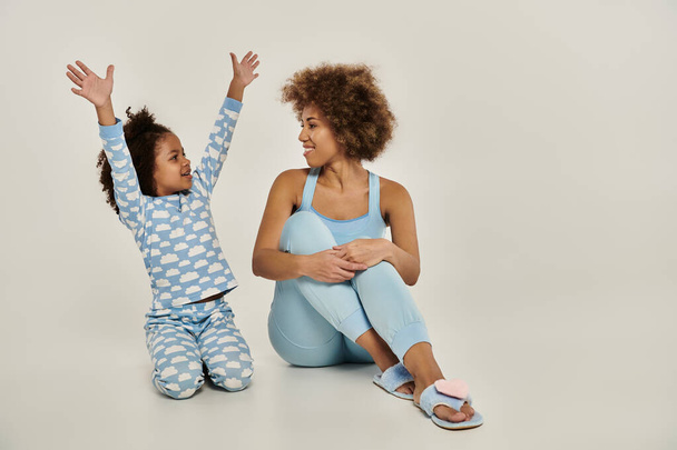Una madre y una hija afroamericanas extáticas en pijama sentadas en el suelo, brazos levantados al cielo al unísono. - Foto, imagen