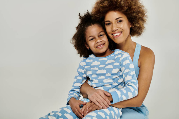 Alegre madre e hija afroamericana abrazándose firmemente en un acogedor pijama sobre un fondo gris. - Foto, imagen